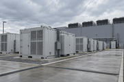 Synthetic editorial illustration. An industrial energy-storage yard. Rows of grey ventilated containers stand on concrete pads beside a windowless building with rooftop cooling units under an overcast sky.