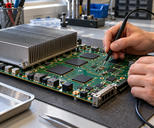 Synthetic editorial illustration. Hands connect a black probe to a large green circuit board on a laboratory bench. A broad finned metal heat sink, blank black chips, traces and sockets fill the board in soft daylight.