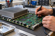 Synthetic editorial illustration. Hands connect a black probe to a large green circuit board on a laboratory bench. A broad finned metal heat sink, blank black chips, traces and sockets fill the board in soft daylight.