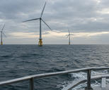 Synthetic editorial illustration. Generic utility wind turbines operating in open water, viewed from a service-vessel railing. Offshore wind electricity production, not oil or gas extraction. No actual named company, product, site, person or event is depicted.