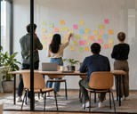 Synthetic editorial illustration. Four people seen from behind discuss blank pastel sticky notes on a meeting-room wall. A plain laptop back and closed device sit on a wooden table inside the daylit glass room.