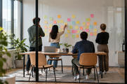 Synthetic editorial illustration. Four people seen from behind discuss blank pastel sticky notes on a meeting-room wall. A plain laptop back and closed device sit on a wooden table inside the daylit glass room.