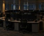 Synthetic editorial illustration. An empty office floor contains rows of dark monitors, phones and tucked-in chairs. Sparse warm lamps and distant city windows provide the only light.