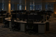 Synthetic editorial illustration. An empty office floor contains rows of dark monitors, phones and tucked-in chairs. Sparse warm lamps and distant city windows provide the only light.