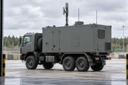 Synthetic editorial illustration. One stationary truck-mounted enclosed electronics shelter with a small communications mast, viewed through a maintenance-bay opening.