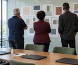 Synthetic editorial illustration. Three people of different builds viewed from behind review paper colour-block reports pinned in a glass meeting room. Plain closed dark laptops, a blank notebook and water glass rest on the table in daylight.