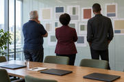 Synthetic editorial illustration. Three people of different builds viewed from behind review paper colour-block reports pinned in a glass meeting room. Plain closed dark laptops, a blank notebook and water glass rest on the table in daylight.