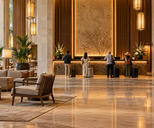 Synthetic editorial illustration. A hotel lobby. Four rear-facing guests with wheeled suitcases stand at a warmly lit stone reception desk, with lounge chairs, plants and a loaded brass luggage trolley across the polished floor.