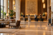 Synthetic editorial illustration. A hotel lobby. Four rear-facing guests with wheeled suitcases stand at a warmly lit stone reception desk, with lounge chairs, plants and a loaded brass luggage trolley across the polished floor.