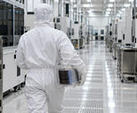 Synthetic editorial illustration. A hooded technician seen from behind carries a transparent cassette containing a stack of reflective wafers down a bright cleanroom aisle. Plain white equipment and stainless carts line the reflective floor.