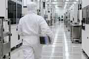 Synthetic editorial illustration. A hooded technician seen from behind carries a transparent cassette containing a stack of reflective wafers down a bright cleanroom aisle. Plain white equipment and stainless carts line the reflective floor.