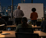 Synthetic editorial illustration. An operations office. Rear-facing workers sit at desks with dark laptops while two colleagues stand beside a whiteboard of empty connected boxes; desk lamps glow against city windows at night.