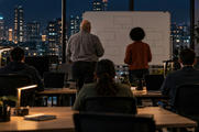 Synthetic editorial illustration. An operations office. Rear-facing workers sit at desks with dark laptops while two colleagues stand beside a whiteboard of empty connected boxes; desk lamps glow against city windows at night.