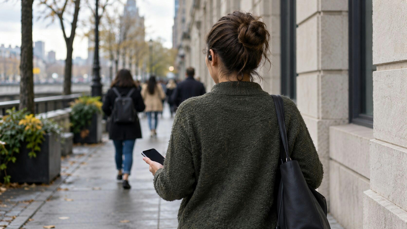 Synthetic editorial illustration. A person viewed from behind walks along a city pavement wearing a small dark earbud and holding a phone with a dark screen. Other pedestrians walk away ahead beside plain buildings and planters.