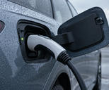 Synthetic editorial illustration. An electric-vehicle charging connector is seated in an open vehicle port, with raindrops, a connected cable and a small green status light.