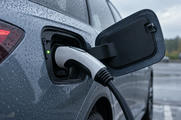 Synthetic editorial illustration. An electric-vehicle charging connector is seated in an open vehicle port, with raindrops, a connected cable and a small green status light.