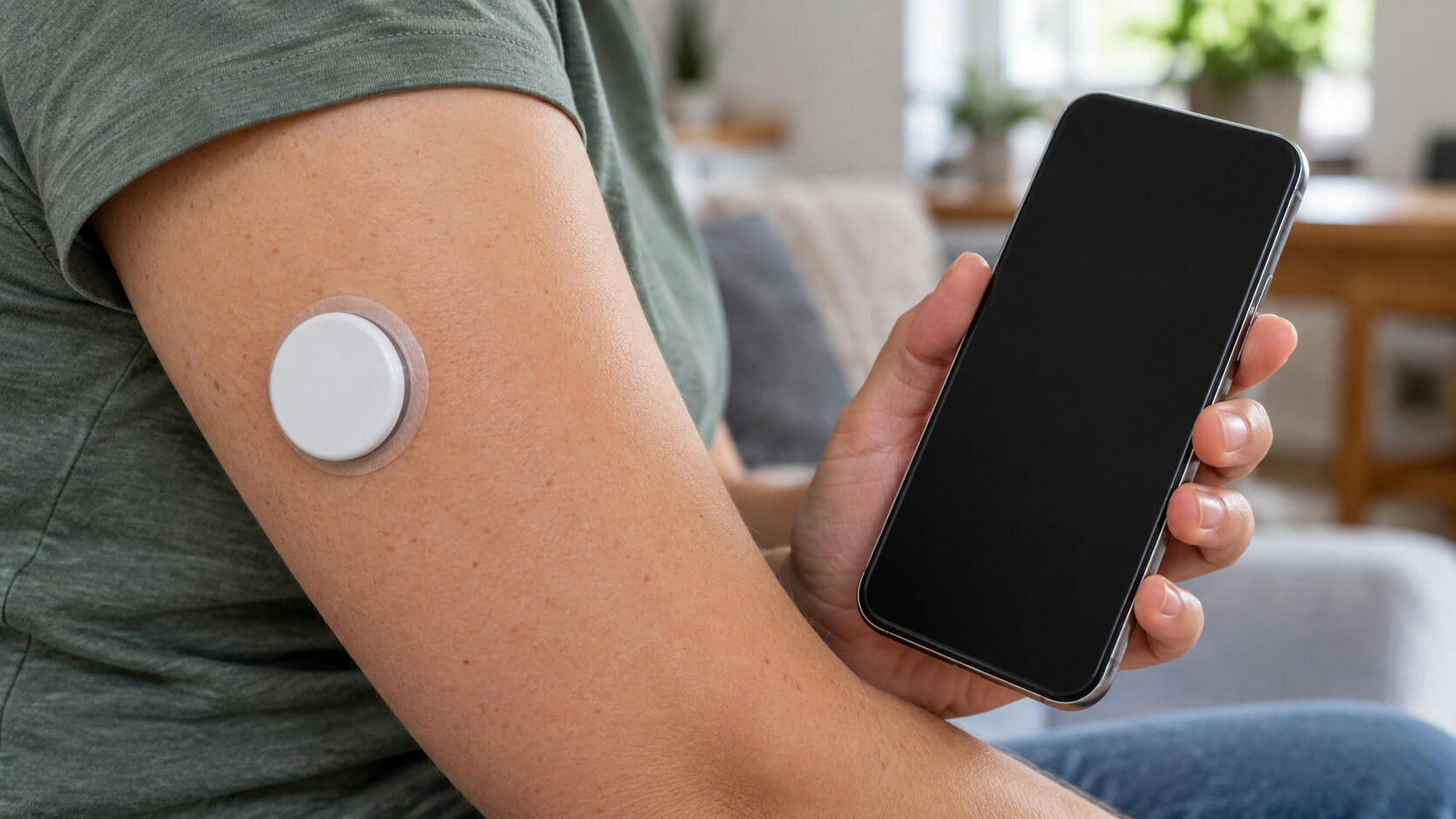 Synthetic editorial illustration. A plain round white sensor sits on an upper arm with a transparent adhesive edge. The person holds a phone with a completely dark screen against a softly blurred domestic background.