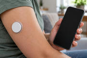 Synthetic editorial illustration. A plain round white sensor sits on an upper arm with a transparent adhesive edge. The person holds a phone with a completely dark screen against a softly blurred domestic background.
