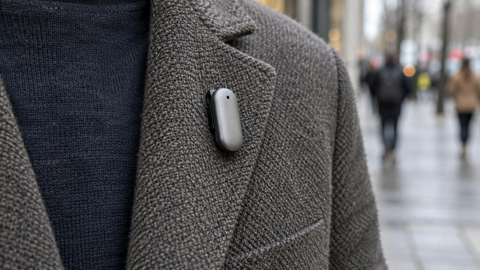 Synthetic editorial illustration. A small plain silver device with a single dark opening clips to the lapel of a textured grey jacket. The close crop excludes the wearer's head, while an urban pavement and distant walkers blur behind.
