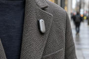 Synthetic editorial illustration. A small plain silver device with a single dark opening clips to the lapel of a textured grey jacket. The close crop excludes the wearer's head, while an urban pavement and distant walkers blur behind.