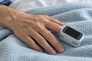 Synthetic editorial illustration. A plain white-and-grey clip device with a completely dark display sits on a patient's finger. The hand rests on a pale blue textured hospital blanket in soft clinical light.