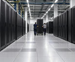 Synthetic editorial illustration. A data-centre hall contains long rows of black server cabinets. Two rear-facing technicians wheel a tall rack along the raised-floor aisle beneath cable trays.