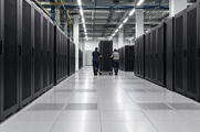 Synthetic editorial illustration. A data-centre hall contains long rows of black server cabinets. Two rear-facing technicians wheel a tall rack along the raised-floor aisle beneath cable trays.