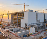 Synthetic editorial illustration. A huge white industrial building under construction rises above exposed steel framing, pipes and service tanks. Cranes and small vehicles occupy the site while low morning sunlight touches the facade.
