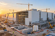Synthetic editorial illustration. A huge white industrial building under construction rises above exposed steel framing, pipes and service tanks. Cranes and small vehicles occupy the site while low morning sunlight touches the facade.