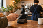 Synthetic editorial illustration. A cafe counter. A hand holds a phone with its plain back toward the camera near a dark payment terminal, while a barista seen from behind works at a coffee machine.