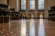 Synthetic editorial illustration. An exchange hall contains curved trading desks and dark display panels. Small paper confetti pieces lie across the polished floor as three distant rear-facing people walk toward the doors.