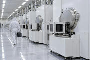 Synthetic editorial illustration. A semiconductor cleanroom. An engineer in a hooded white suit seen from behind carries a closed dark case along a row of white process machines with circular metal chambers and wafer load ports.