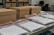 Synthetic editorial illustration. An archive room contains metal shelving and a worktable. Plain brown boxes and sealed transparent bags of blank documents sit in the foreground, with a dark monitor and empty chair behind.