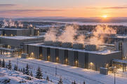 Synthetic editorial illustration. Low industrial compute buildings and rows of cooling units stand amid snow and conifer forest at dawn. Pale vapour rises above the roofs as warm sunrise light touches pipes and service roads.