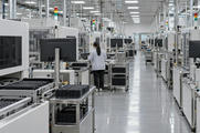 Synthetic editorial illustration. A semiconductor testing hall. An engineer seen from behind checks a black component tray among long rows of enclosed test machines, tray-loading rails and monitors facing away.