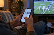 Synthetic editorial illustration. An adult uses a complete powered smartphone in a modest evening living room while a softly defocused television shows a generic American-football game in a separate deeper plane.