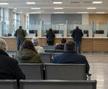 Synthetic editorial illustration. A public-service office contains a glass-partitioned counter and rows of waiting seats. Rear-facing visitors sit or stand nearby, beside a ticket machine with a blank dark display.
