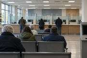 Synthetic editorial illustration. A public-service office contains a glass-partitioned counter and rows of waiting seats. Rear-facing visitors sit or stand nearby, beside a ticket machine with a blank dark display.
