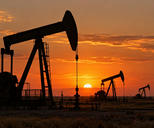 Synthetic editorial illustration. An oilfield. Several pumpjacks stand in dark silhouette across a flat scrub-covered landscape, with low pipework and fencing beneath an orange sunset sky.