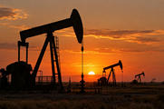 Synthetic editorial illustration. An oilfield. Several pumpjacks stand in dark silhouette across a flat scrub-covered landscape, with low pipework and fencing beneath an orange sunset sky.