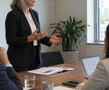 Synthetic editorial illustration. A standing adult addresses two seated colleagues around an occupied meeting table with a populated meeting pack.
