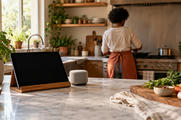 Synthetic editorial illustration. A home kitchen. A dark tablet on a wooden stand and a small fabric-covered speaker sit on the island beside vegetables, while a rear-facing person in a rust apron cooks at the stove.