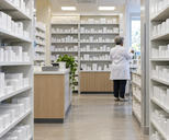 Synthetic editorial illustration. A pharmacy aisle is lined with shelves of mostly plain white boxes and bottles. A rear-facing grey-haired pharmacist in a white coat checks a small carton near the bright window.