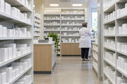 Synthetic editorial illustration. A pharmacy aisle is lined with shelves of mostly plain white boxes and bottles. A rear-facing grey-haired pharmacist in a white coat checks a small carton near the bright window.