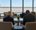Synthetic editorial illustration. An airport business lounge. Two advisers viewed from behind sit together looking down at closed white folders on a low table, with empty apron visible through broad windows.