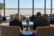 Synthetic editorial illustration. An airport business lounge. Two advisers viewed from behind sit together looking down at closed white folders on a low table, with empty apron visible through broad windows.