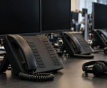 Synthetic editorial illustration. A trading desk contains multi-line phones, headsets and switched-off monitors. A close view emphasizes an unmarked black handset, blank buttons and coiled cable, with rear-facing workers blurred in the background.