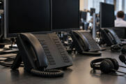 Synthetic editorial illustration. A trading desk contains multi-line phones, headsets and switched-off monitors. A close view emphasizes an unmarked black handset, blank buttons and coiled cable, with rear-facing workers blurred in the background.