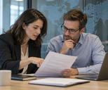 Synthetic editorial illustration. Two adults discuss a populated report beside a subtle dotted world-map glass partition and complete laptop.