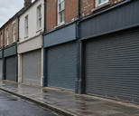 Synthetic editorial illustration. A row of brick and plaster shopfronts has closed metal shutters and blank fascias. The empty pavement and wet road recede under flat grey morning light.